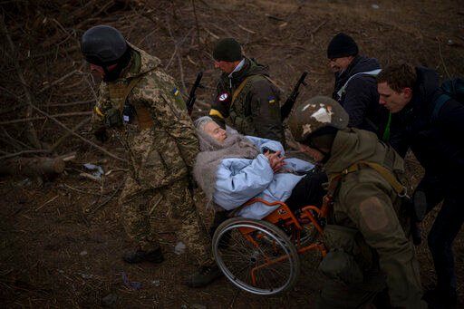 Ukrainian soldiers and militiamen carry a woman in a wheelchair as the artillery echoes nearby, while people flee Irpin on the outskirts of Kyiv, Ukraine, Monday, March 7, 2022. Russia announced yet another cease-fire and a handful of humanitarian corridors to allow civilians to flee Ukraine. Previous such measures have fallen apart and Moscow