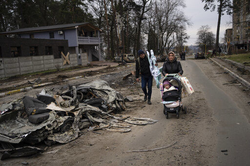 A family walks pass a car crushed by a Russian tank in Bucha, in the outskirts of Kyiv, Ukraine, Tuesday, April 5, 2022. Ukraine