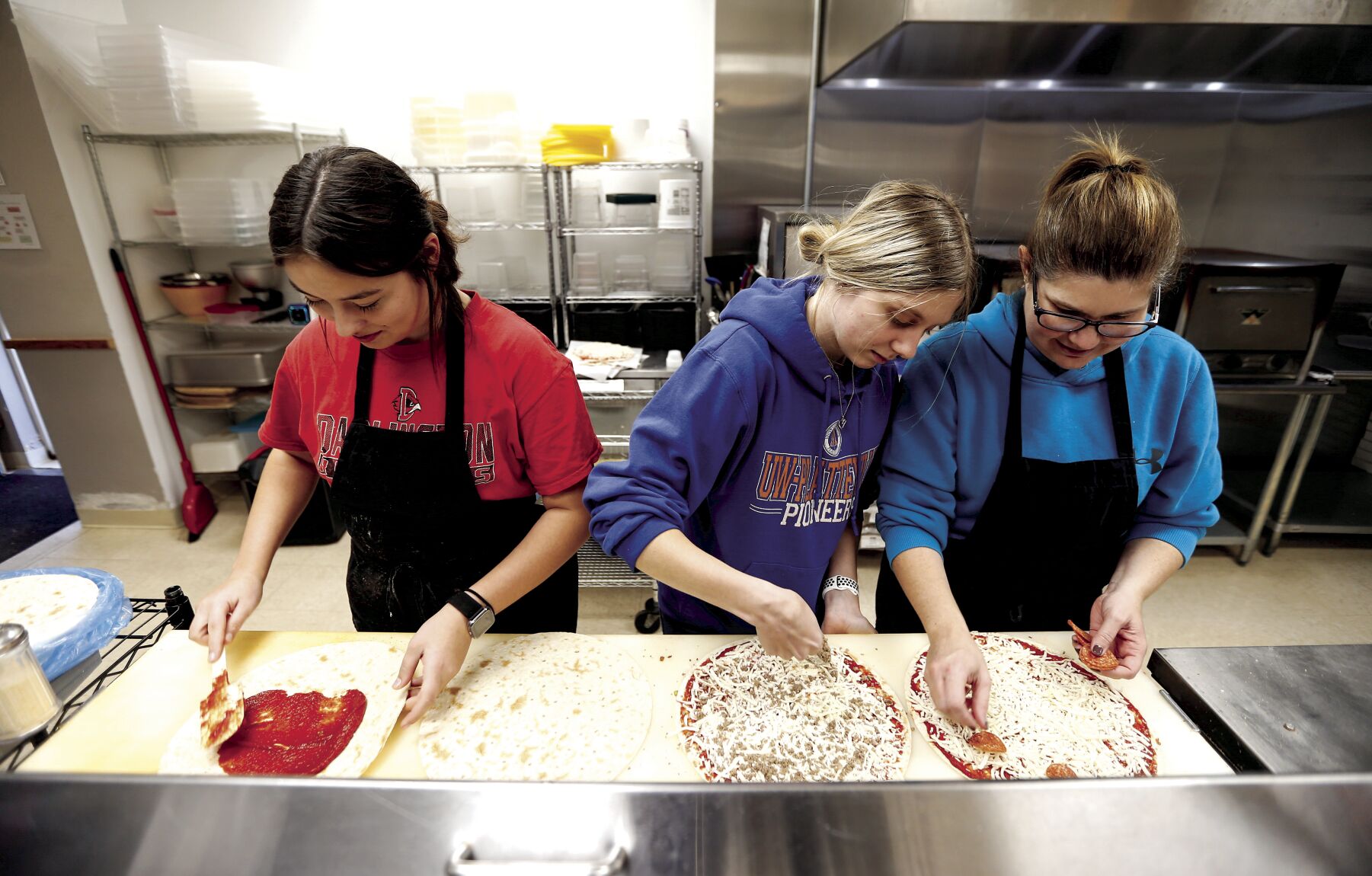 Alexis Mosley (left), 16, works with Ava Smith and her mother, co-owner Val Smith, on making pizzas at Vinger’s Pizza in Darlington, Wis., before opening on Saturday.    PHOTO CREDIT: Dave Kettering