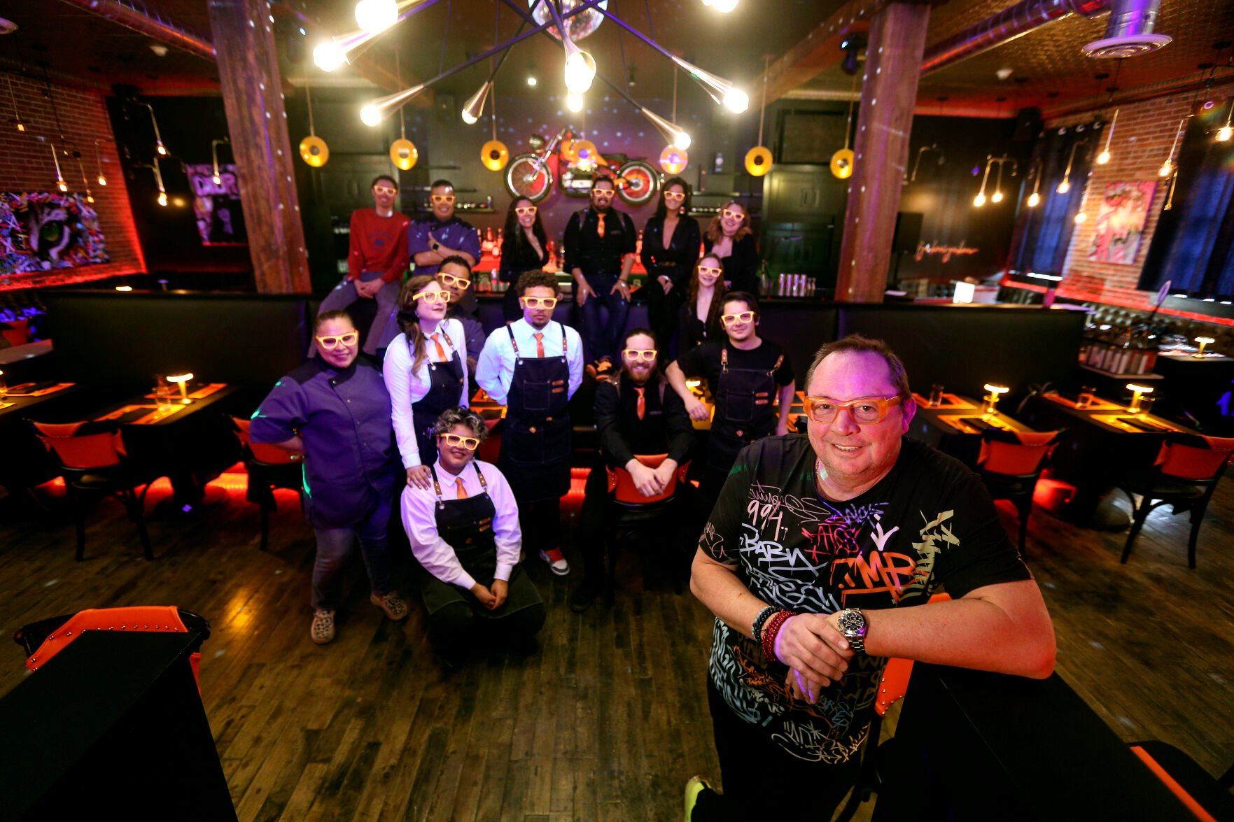 Eric Bonnetain (right), owner of Bread & Vine, will open a Dubuque location in the Millwork District today. He is pictured with his staff — mimicking his signature orange glasses — at the restaurant and entertainment space.    PHOTO CREDIT: Dave Kettering