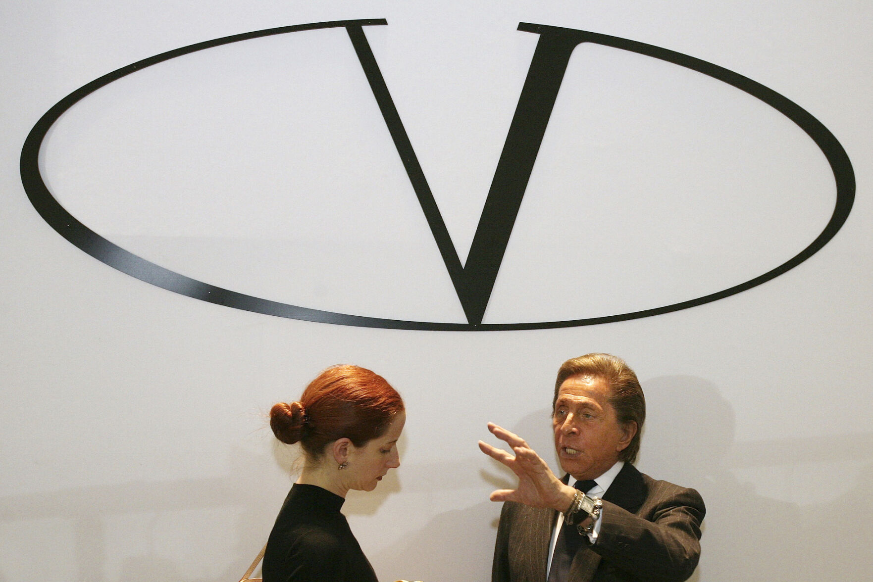 <p>FILE - Italian fashion designer Valentino, right, standing underneath the logo of his fashion house, answers the questions of a fashion reporter prior to the presentation of his Haute Couture Spring-Summer 2008 fashion collection, on Jan. 23, 2008, in Paris. French luxury conglomerate Kering has reached a cash deal to purchase a 30% stake in Italian fashion house Valentino for 1.7 billion euros from a Qatari investment firm. (AP Photo/Thibault Camus, File)</p>   PHOTO CREDIT: THIBAULT CAMUS