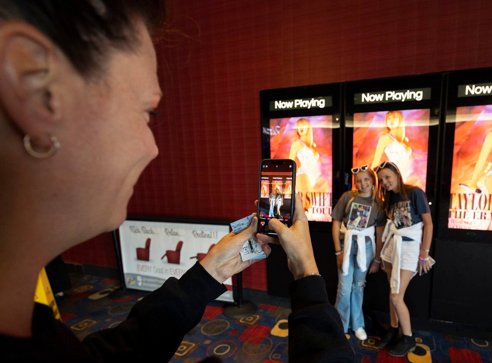 Lindsey Behnke takes a photo of Nora Reuter, 10, (left) and Hannah Behnke, 11, on Saturday in front of movie posters promoting the “Taylor Swift: The Eras Tour” concert film at Phoenix Theatres in Dubuque.    PHOTO CREDIT: Stephen Gassman